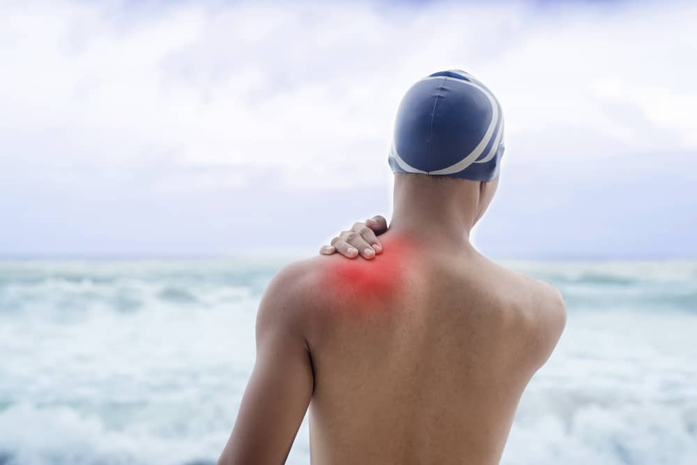 A swimmer stands on the beach grabbing his shoulder due to back pain from swimming in the ocean.